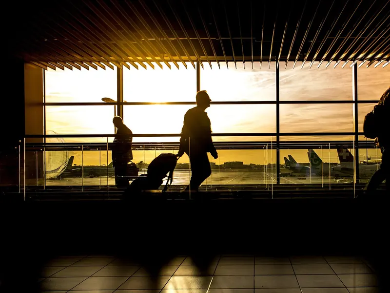 Travellers with luggage silhouetted against an airport window at sunrise.