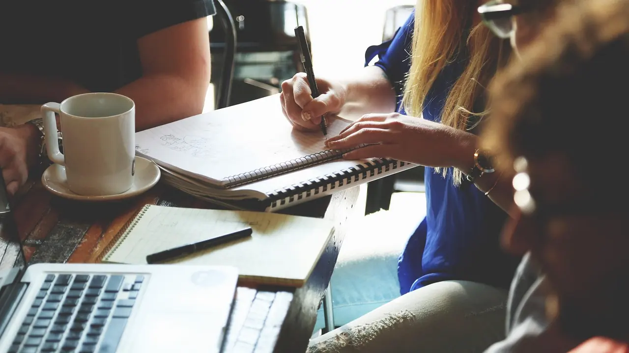 Two people working through paperwork together at a desk, notebook and coffee to one side.